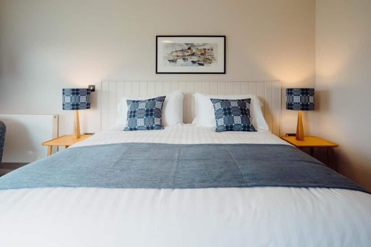 Cama bem arrumada com cabeceira branca, dois travesseiros, manta azul e duas luminárias de mesa com cúpulas estampadas.