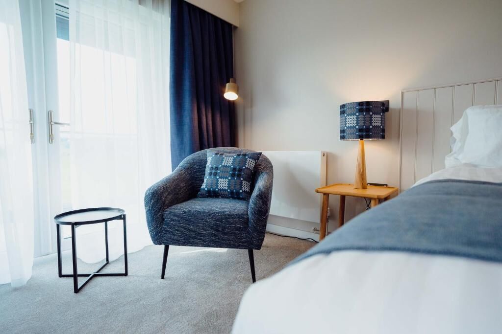 A hotel room with a bed, a chair, a small table, a lamp, and a view of the outdoors.