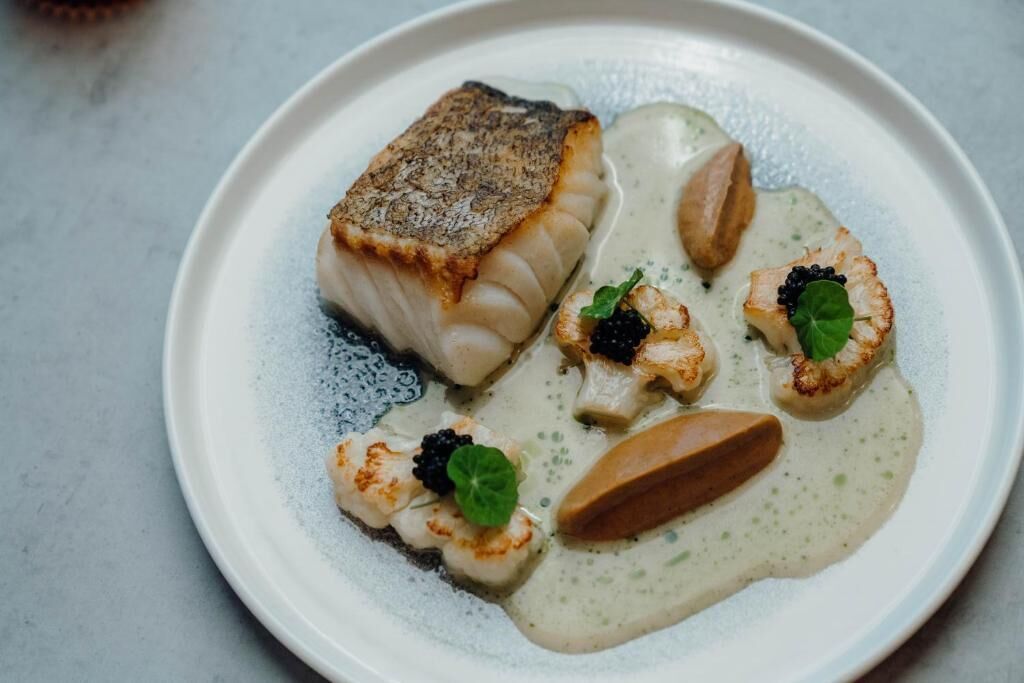 Un plato de comida con un trozo de pescado, vieiras y salsa cremosa.