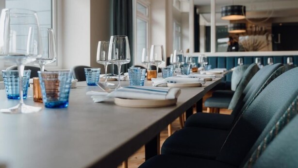 Um restaurante com uma mesa comprida posta para refeição, cadeiras azuis e copos de vidro transparente.
