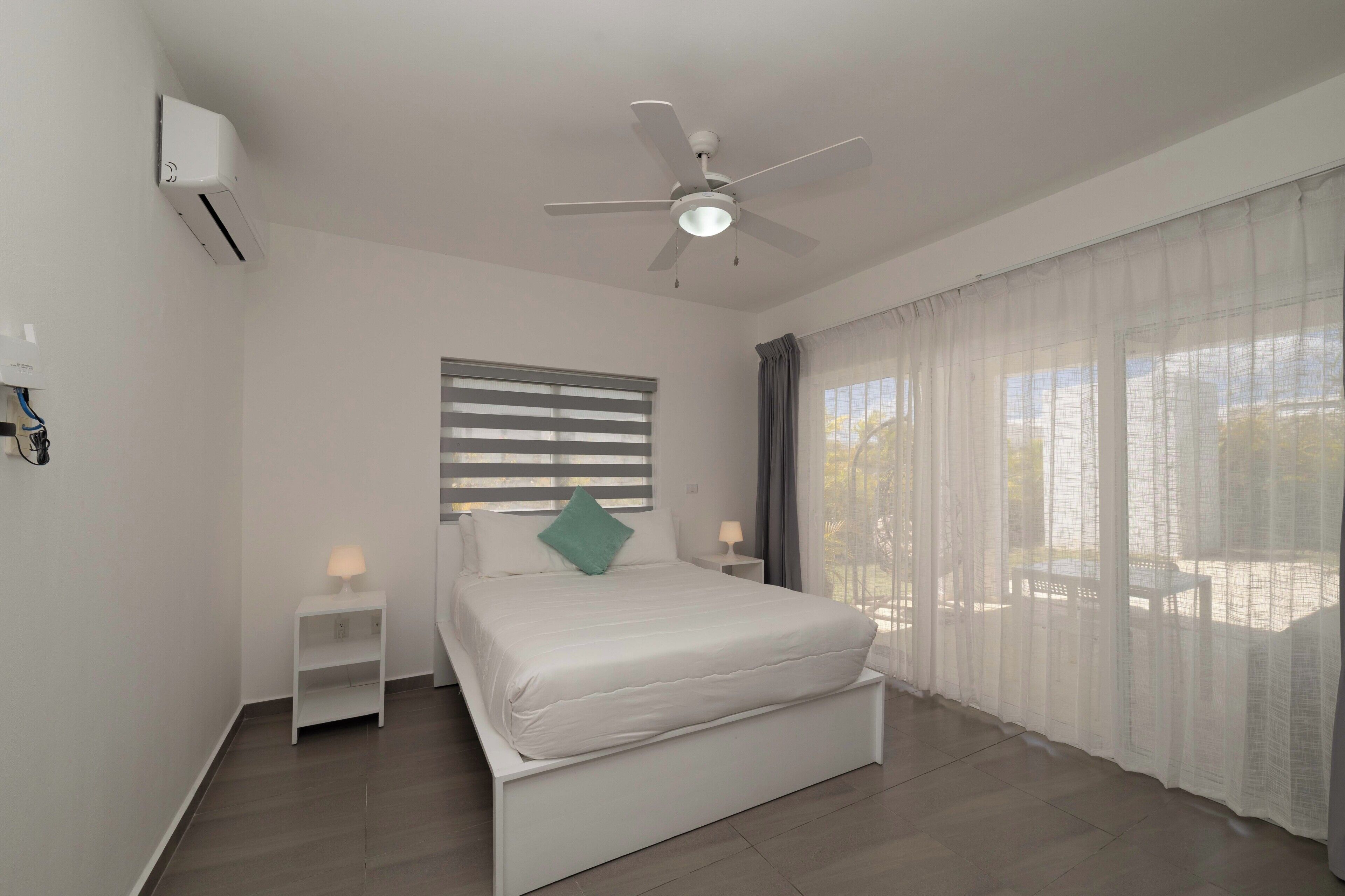 Une chambre à coucher avec un lit, un ventilateur de plafond, une fenêtre avec des rideaux et une petite table de chevet avec une lampe.