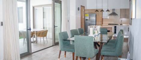 A modern dining area with a glass table and green upholstered chairs, a kitchen with wooden cabinets and stainless steel appliances, and a sliding glass door leading to an outdoor area.