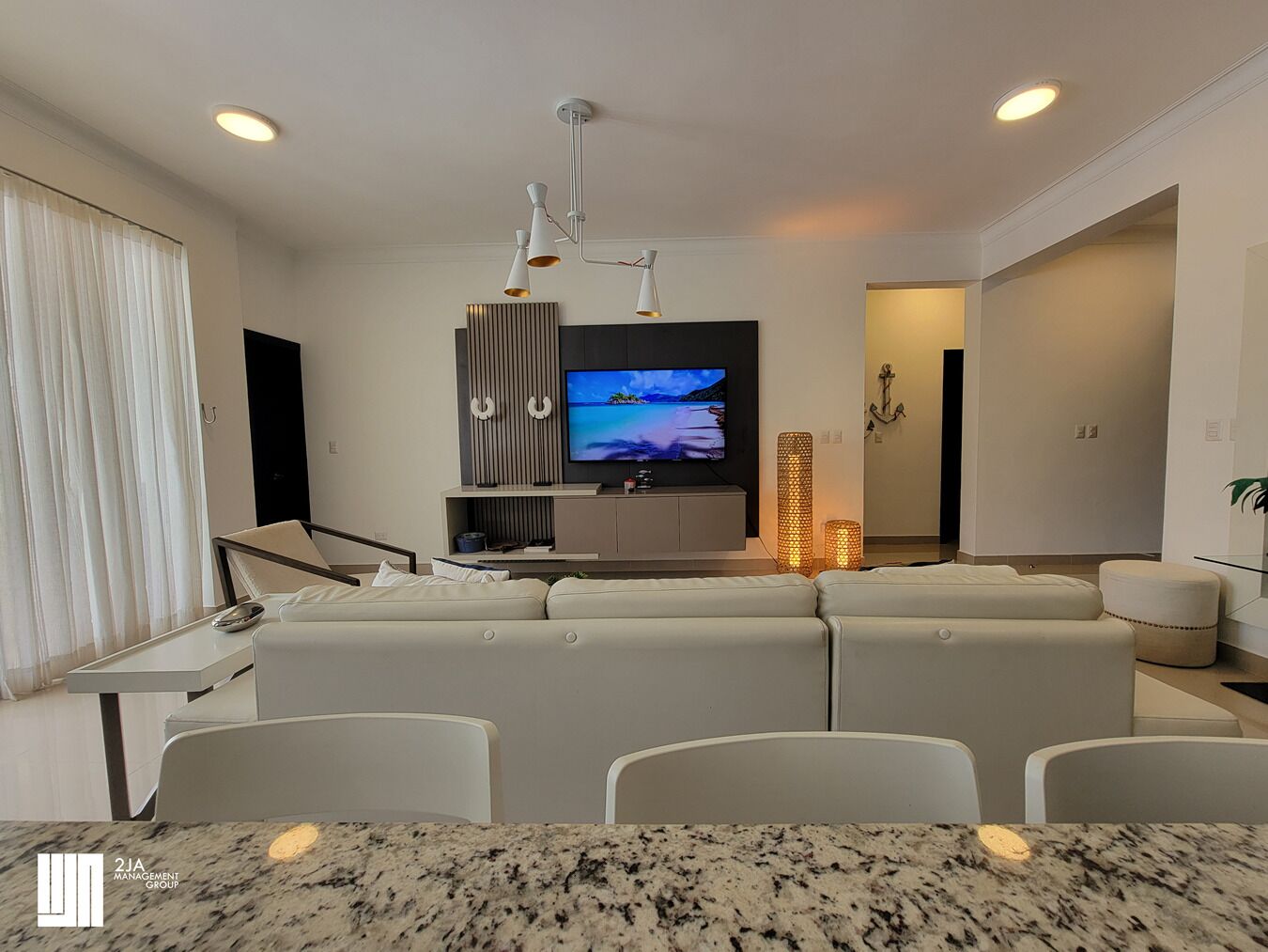 A modern living room with a large TV, white sofas, a dining area with a marble table, and a staircase leading to another level.