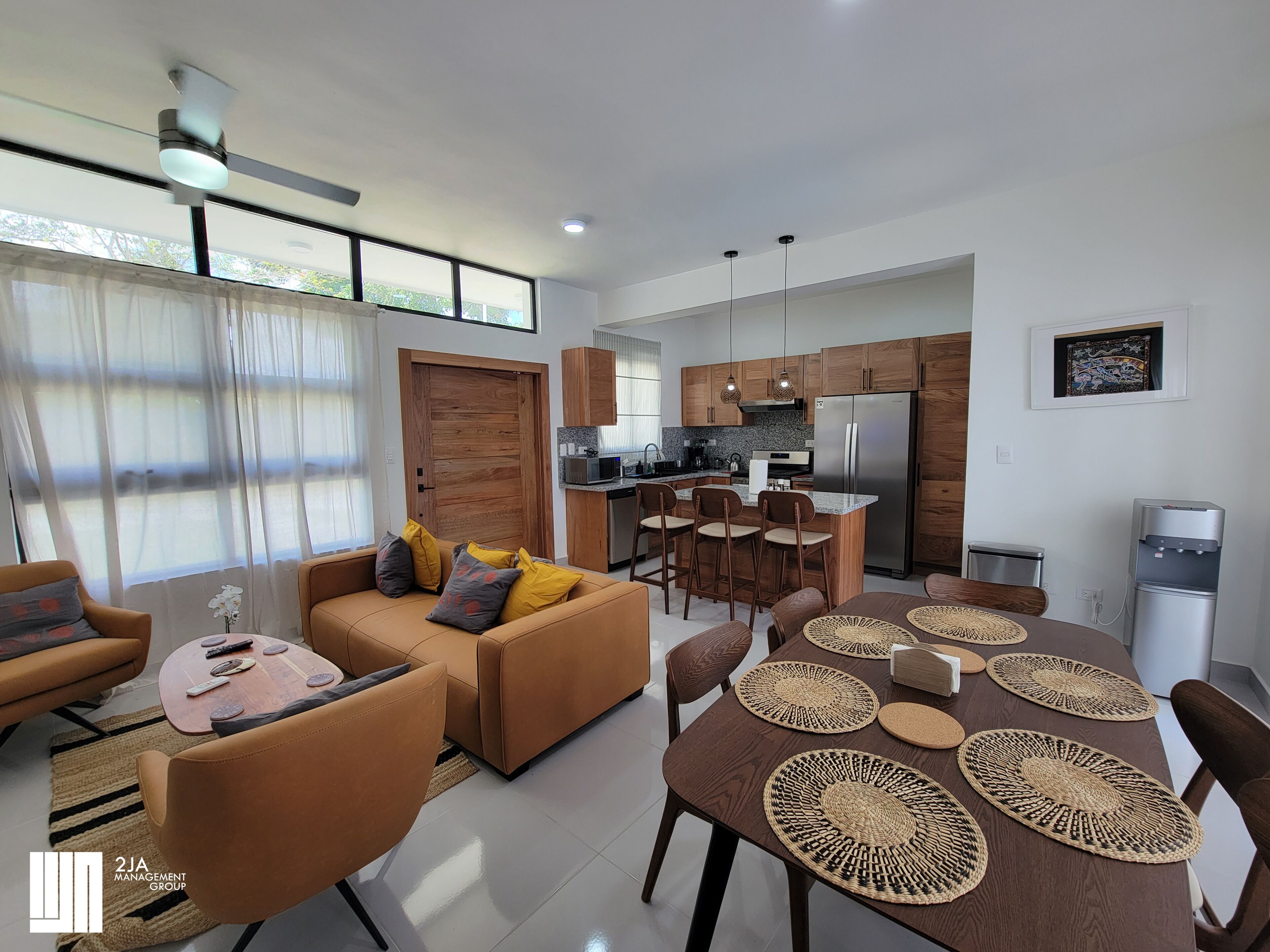 A modern living room with a sofa, chairs, and a dining table with placemats. There is a kitchen area with wooden cabinets and a refrigerator.