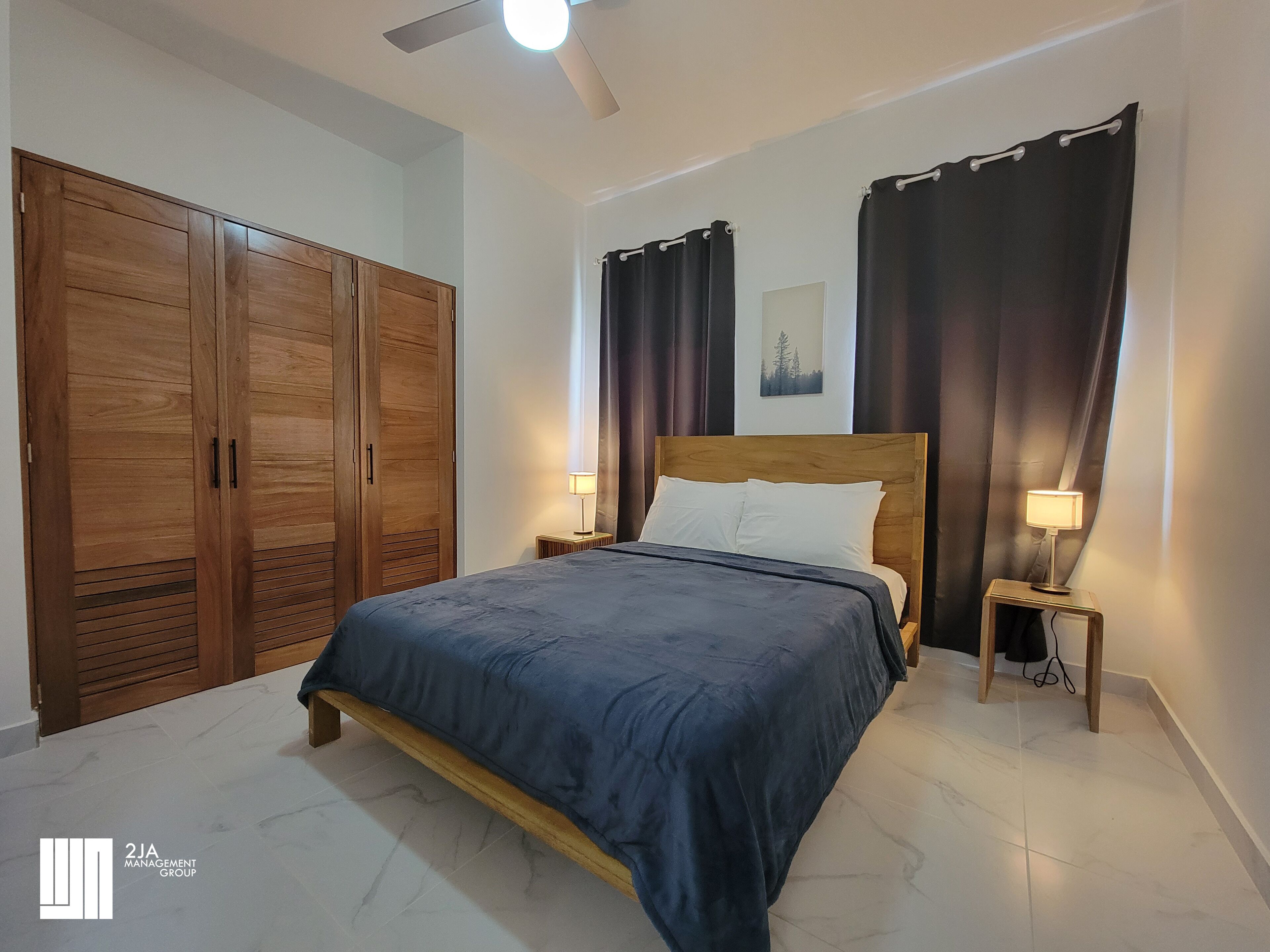 Un dormitorio con una cama grande, armario de madera, dos mesitas de noche con lámparas, cortinas oscuras y un ventilador de techo.