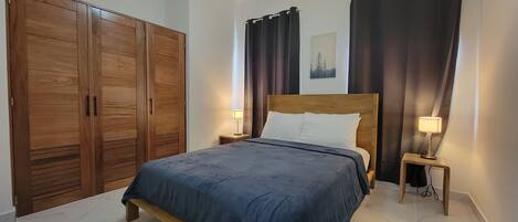 Un dormitorio con una cama grande, armario de madera, dos mesitas de noche con lámparas, cortinas oscuras y un ventilador de techo.