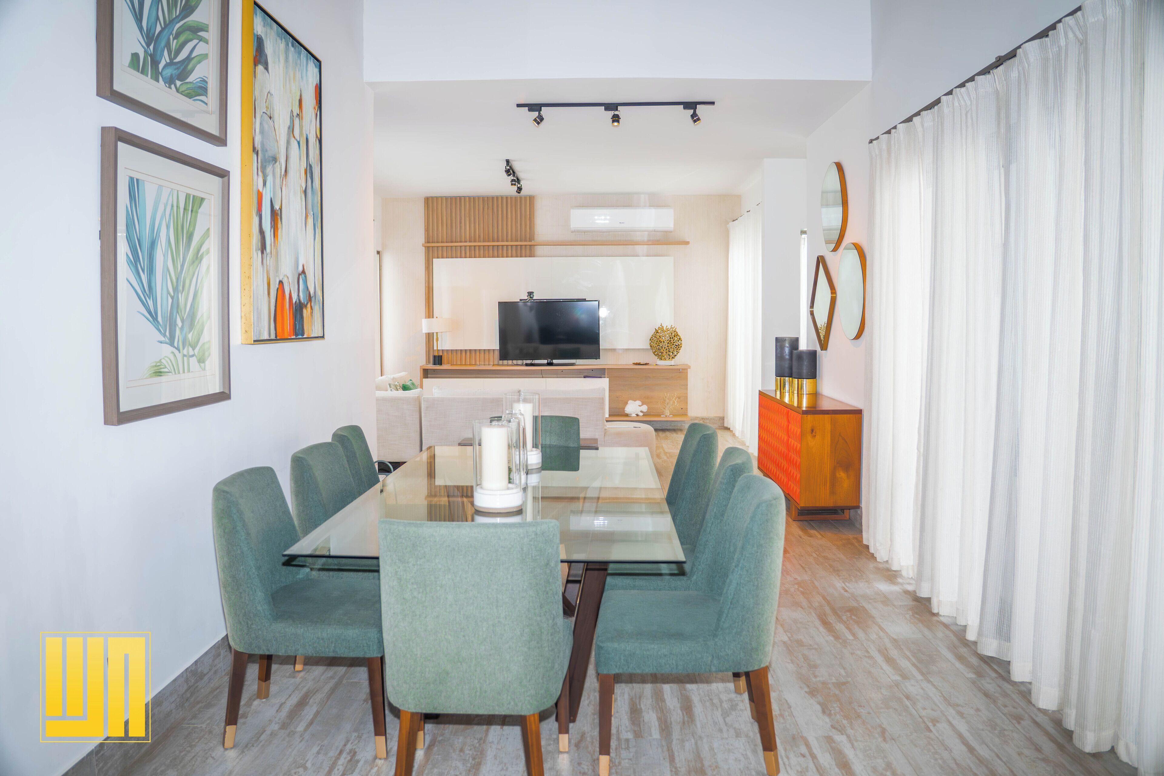 A modern living room with a glass dining table, teal chairs, a flat-screen TV, and a wooden cabinet.
