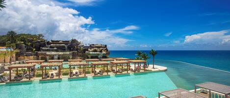 A resort pool with lounge areas, a waterfall, and a view of the ocean.