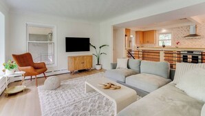 A modern living room with a sofa, armchair, and a TV. There's a kitchen area with wooden cabinets and a stove. - Midcentury Lake Home! (White Bear Lake)