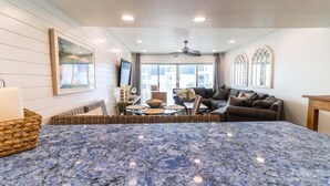 A spacious living room with a large blue marble table, a sofa set, wicker baskets, and a ceiling fan. - Surfside Serenity - Beachfront w/Pool & Ocean View (New Smyrna Beach)