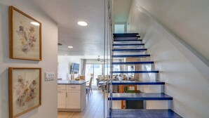 A modern staircase with blue steps leading to a landing, a kitchen area with white cabinets, and framed artwork on the walls. - Surfside Serenity - Beachfront w/Pool & Ocean View (New Smyrna Beach)