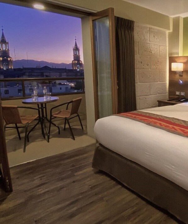 Habitación de hotel con balcón, cama, mesa y sillas, y vistas a dos torres.