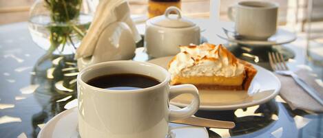 Una mesa con una taza de café, una porción de pastel, un florero con flores y una tetera.