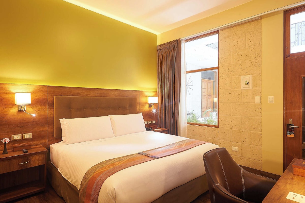 Une chambre d’hôtel avec un grand lit, une tête de lit en bois, deux lampes de chevet, une table de chevet, un bureau et une fenêtre avec des rideaux.