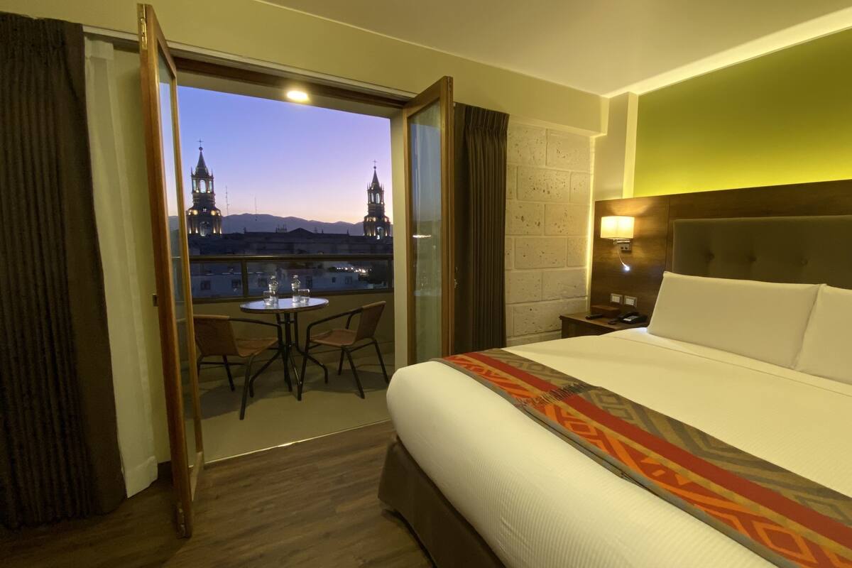 Une chambre d’hôtel avec un grand lit, une petite table avec des chaises, un balcon donnant sur des immeubles et une montagne, et une table de chevet avec une lampe.