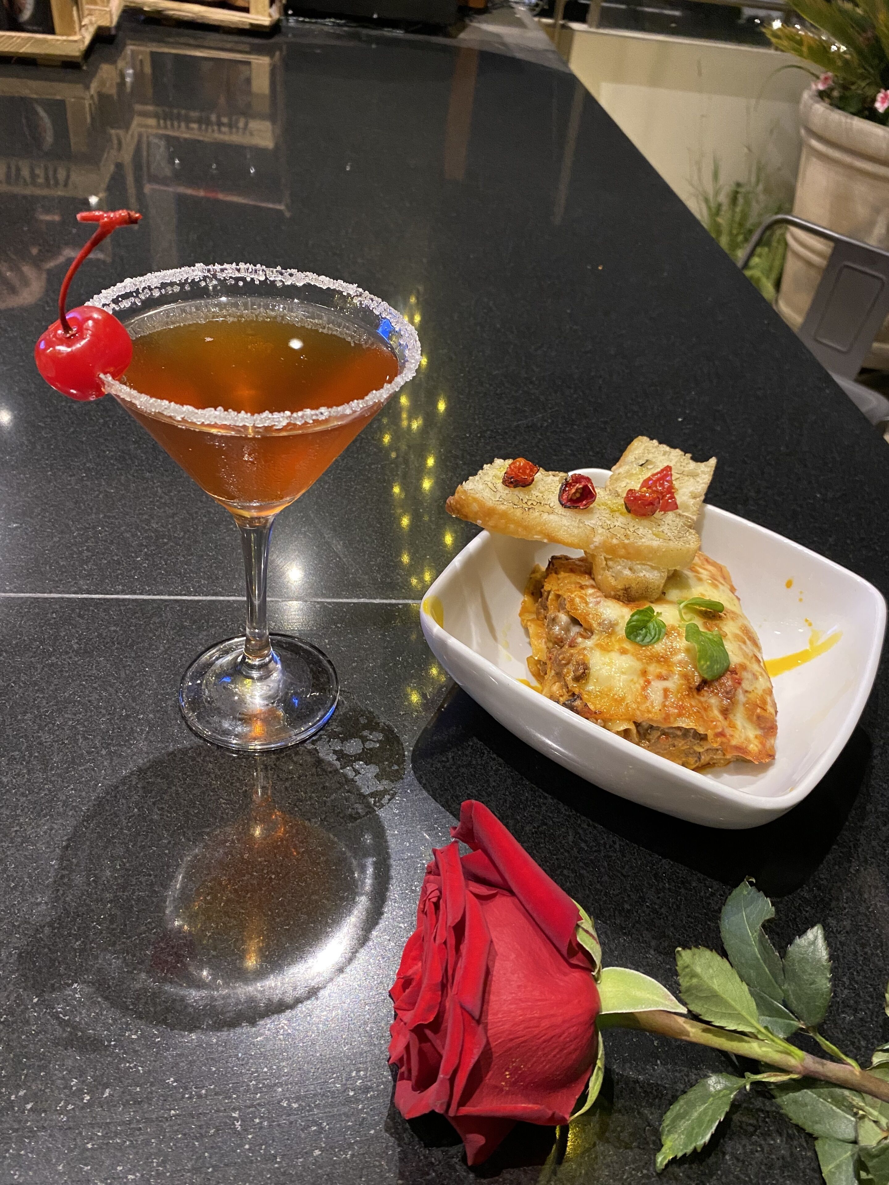 A martini glass with a cherry on the rim, a slice of pizza with toppings, and a red rose on a table.