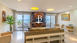 A modern living room with a large window, a sofa, a bench, and a decorative wall piece. - Vista mar 2 suítes e piscina exterior (Florianópolis)