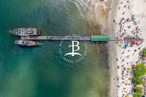 An aerial view of a beach with a pier, boats, and a large 'B' logo.
