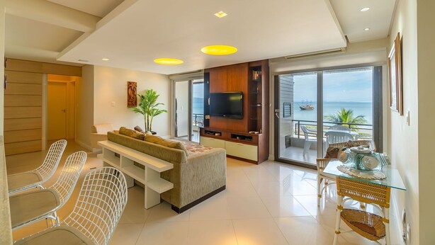 A modern living room with a sofa, chairs, a TV, and a balcony with a view of the sea. - Vista mar 2 suítes e piscina exterior (Florianópolis)