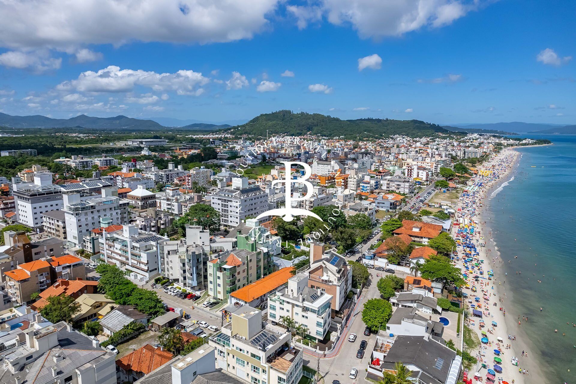 An aerial view of a coastal city with dense urban development, a beach, and a mountainous backdrop.