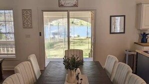 A dining room with a dark wooden table, white upholstered chairs, a chandelier, and a view of a backyard through glass doors. - 5 Bdrm, Amazing Sunsets, deck, flat yard, dock, fish, beach, kayaks... (Sparta)