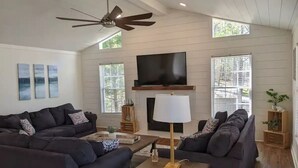 A modern living room with two dark sofas, a wooden coffee table, a floor lamp, a flat-screen TV, and a fireplace. - 5 Bdrm, Amazing Sunsets, deck, flat yard, dock, fish, beach, kayaks... (Sparta)
