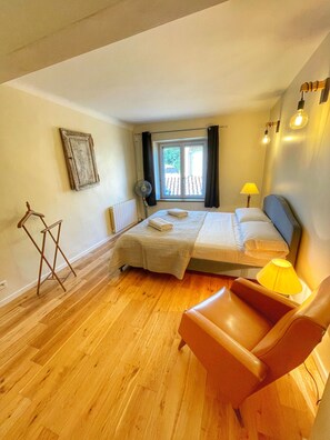 A bedroom with a bed, a chair, a standing fan, a window with curtains, and a lamp.