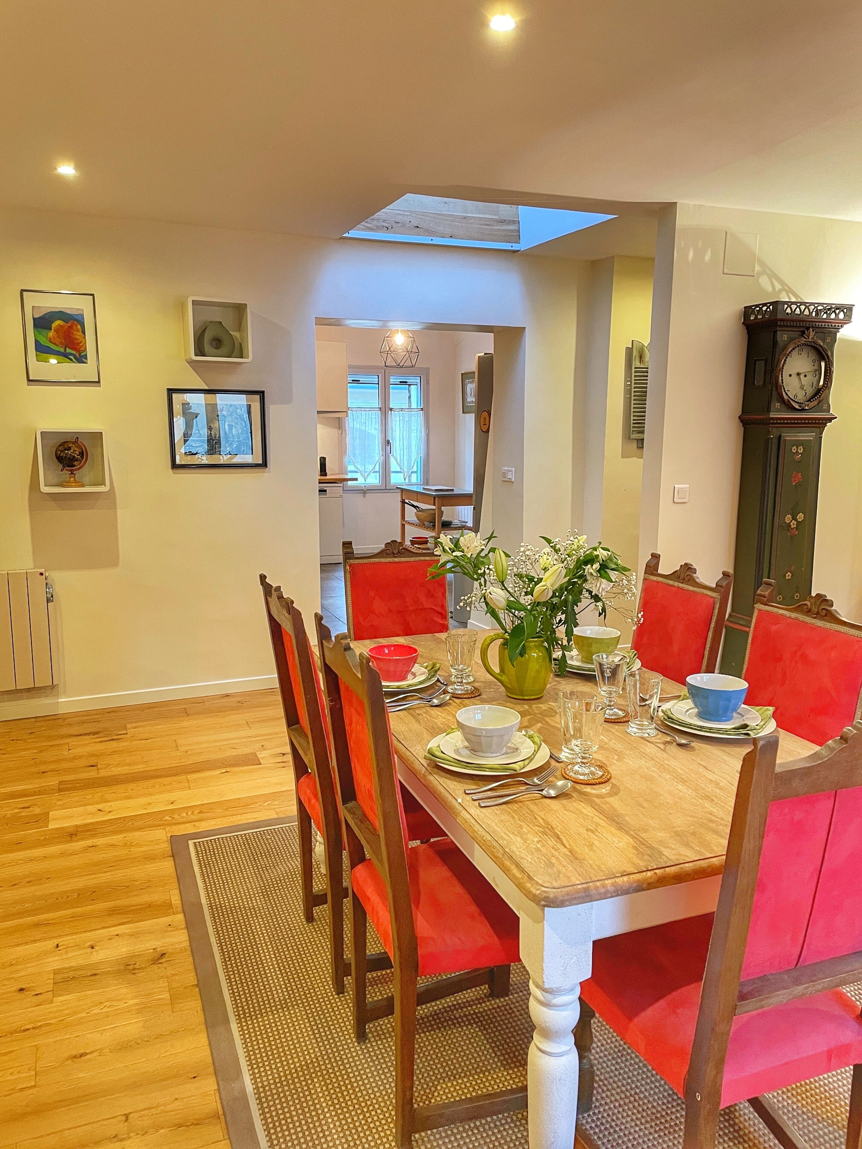Un coin repas avec une table en bois dressée pour six personnes, des chaises à revêtement rouge, un vase avec des fleurs et une horloge fixée au mur.
