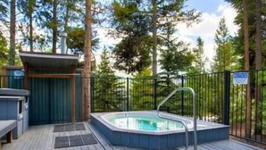 Un jacuzzi en una terraza de madera, rodeado por una valla metálica, con un pequeño edificio y una banca cerca.