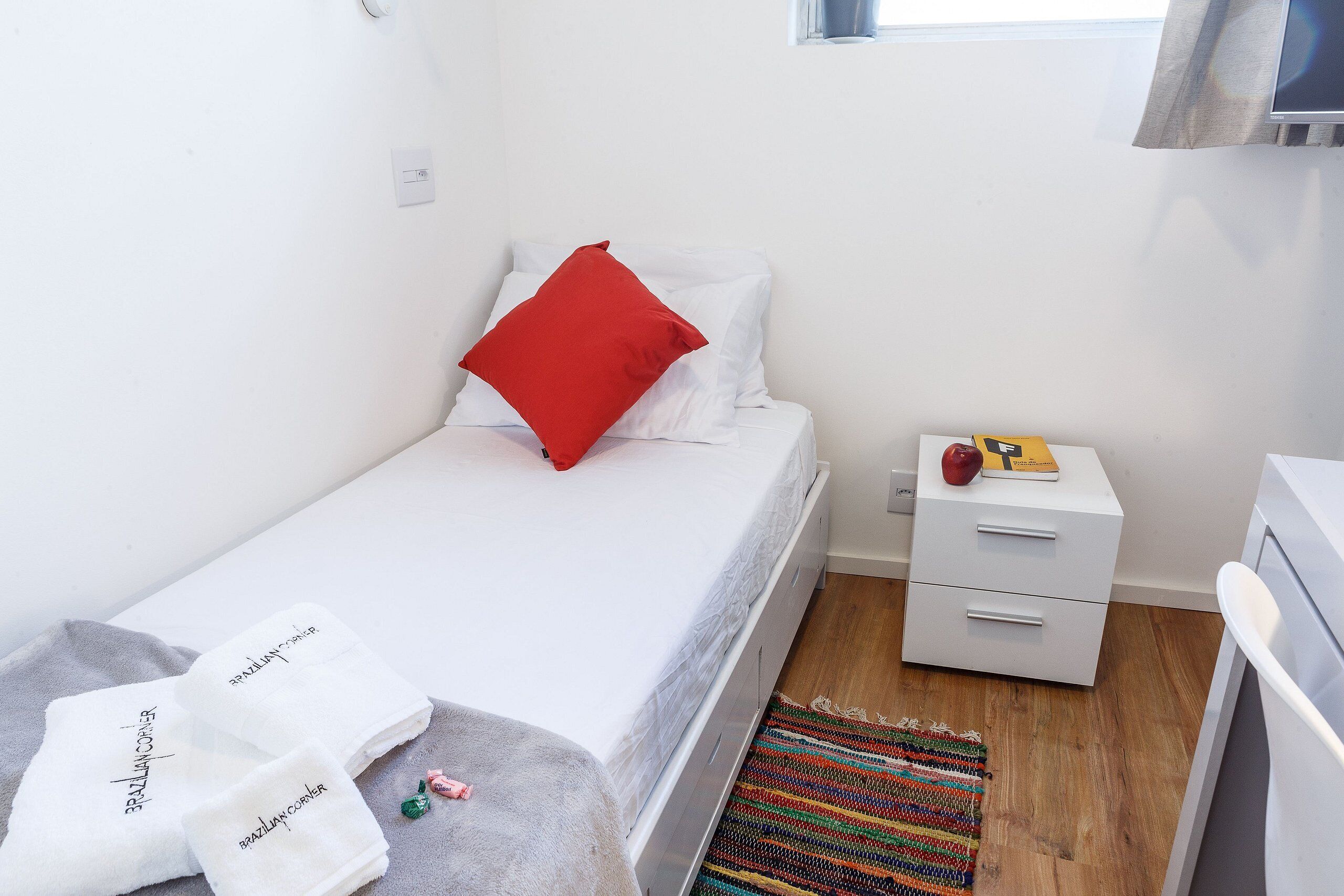 A small, neatly arranged bedroom with a bed, a nightstand, a drawer unit, and a window.