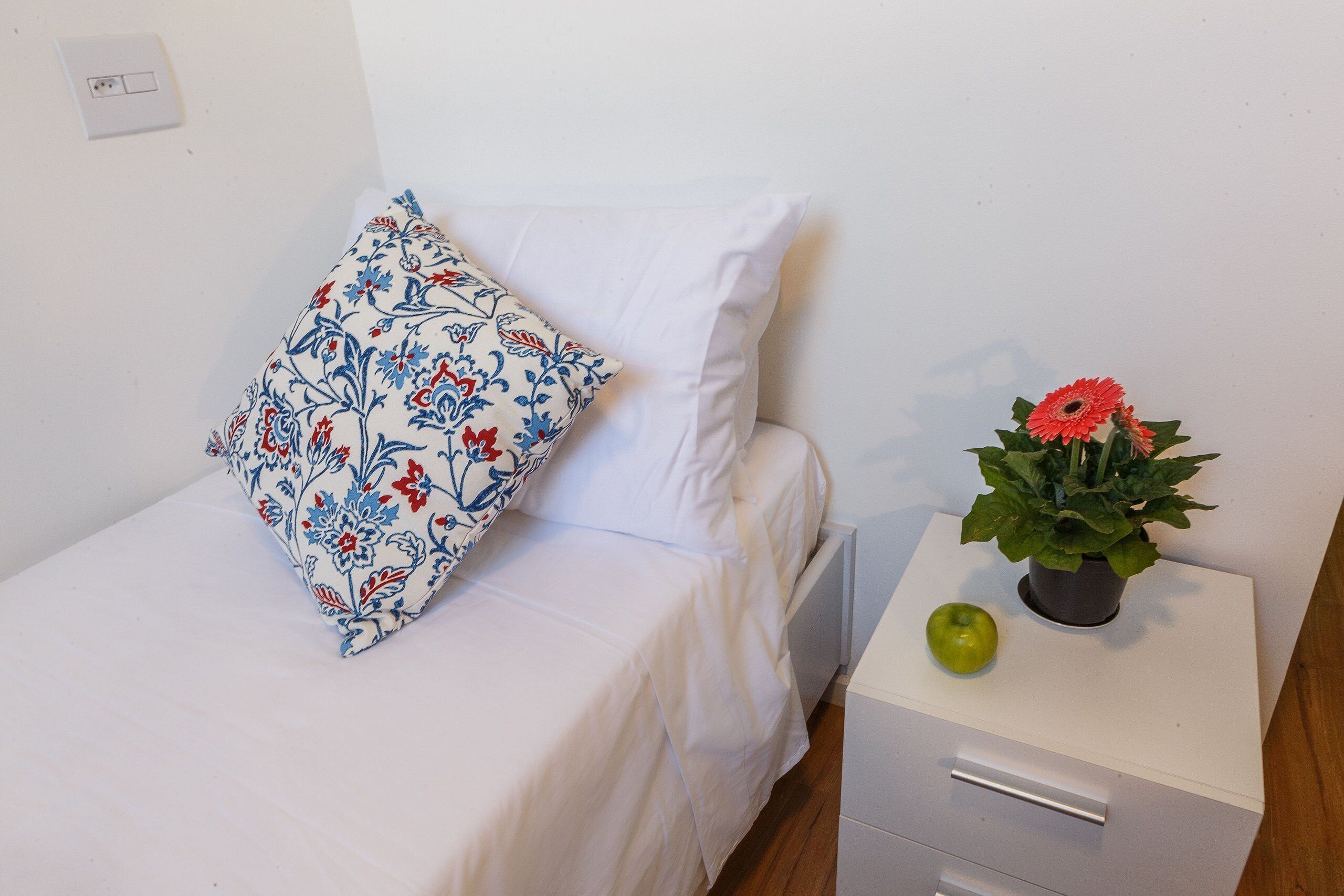 A bed with a white sheet and a floral pillow, a bedside table with a potted plant and an apple, and a white drawer unit.