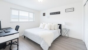 Ein Schlafzimmer mit einem Bett, einem Schreibtisch und einem Stuhl. Zwei Wandbilder und ein Fenster mit Jalousien sind vorhanden.