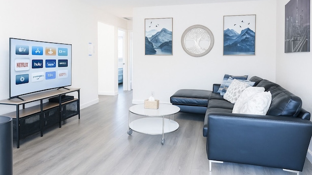Ein modernes Wohnzimmer mit Fernseher, der verschiedene Streaming-Dienste anzeigt, einem dunkelblauen Ledersofa, einem runden Couchtisch und gerahmten Kunstwerken an den Wänden.