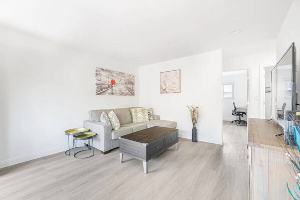 A modern living room with a grey sofa, a wooden coffee table, and two round side tables. There are two framed artworks on the wall and a large mirror on the right. - Comfy 2BR Apt w/ AC/Washer/Dryer/EV - in Sunnyvale (Sunnyvale)