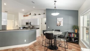 A modern kitchen with a central island, a dining area with chairs, a bookshelf, and a large window. - Inverness Poolside Oasis 3 Bedroom (Inverness)