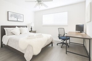 A bedroom with a bed, a desk, a chair, a ceiling fan, a window with blinds, and a framed picture on the wall.