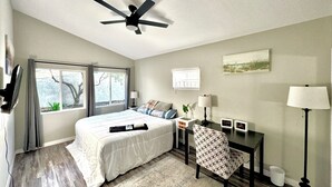 Une chambre à coucher comprenant un lit, un bureau, une chaise, un ventilateur de plafond et deux fenêtres.