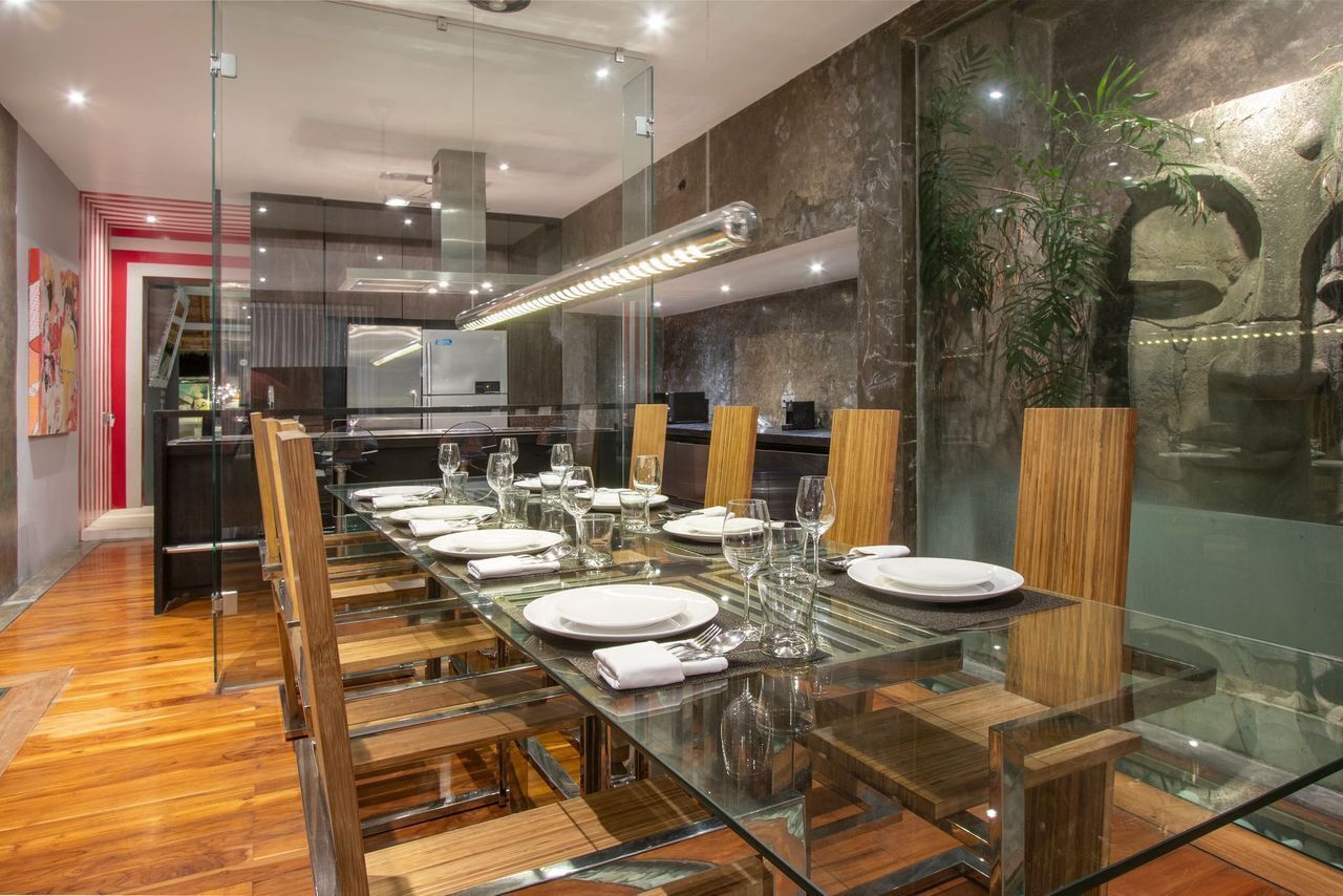 Un espace repas moderne avec une table en verre et des chaises en bois, une cuisine en arrière-plan et un mur décoratif agrémenté de plantes.