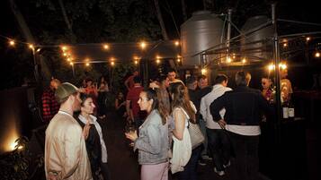 Um grupo de pessoas reunido ao ar livre à noite, possivelmente em um evento social ou festa, com luzes de Natal penduradas e uma grande estrutura industrial ao fundo.
