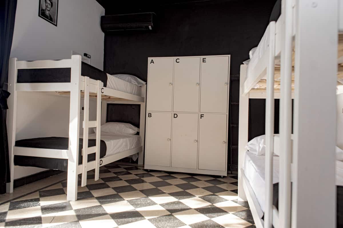 Habitación de residencia estudiantil con literas, suelo a cuadros y un armario blanco con compartimentos etiquetados de la A a la E.