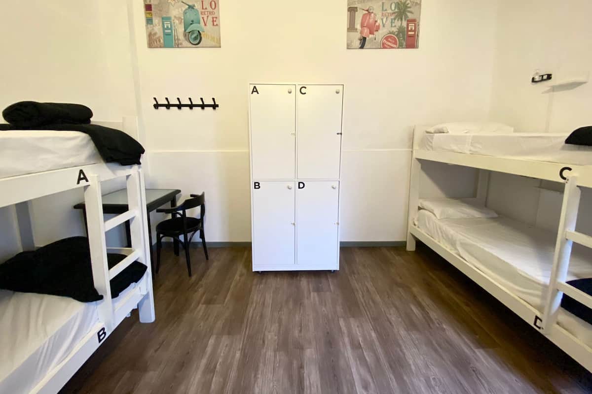 Habitación de residencia con literas identificadas con las letras A, B y C, un armario blanco y dos ganchos en la pared.