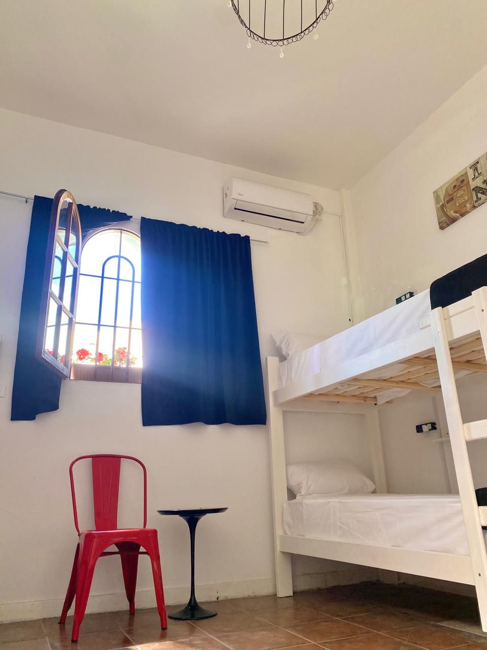 Una habitación pequeña con una litera, una silla roja, una mesita y una ventana con cortinas.