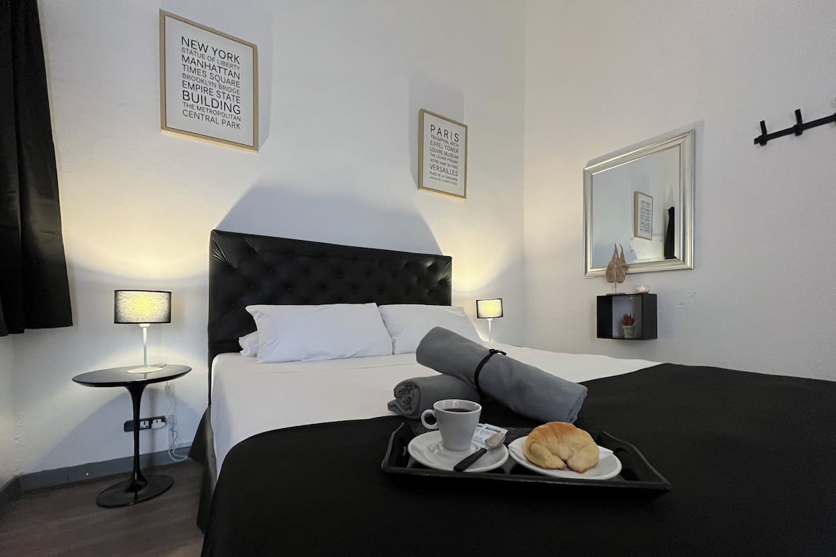 Habitación de hotel con una cama grande, una mesita de noche con lámpara, una bandeja con un croissant y una taza de café, y cuadros en la pared.
