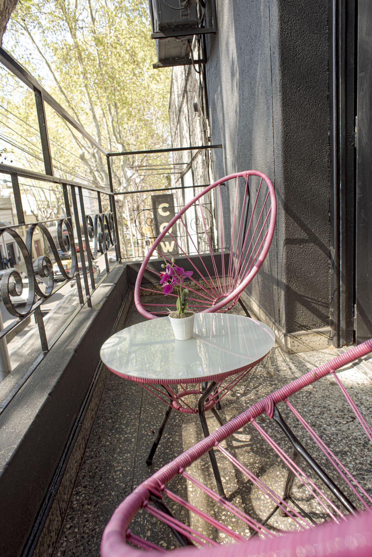 Uma pequena área externa com mesa redonda e uma cadeira rosa, uma orquídea em vaso e um corrimão de metal.