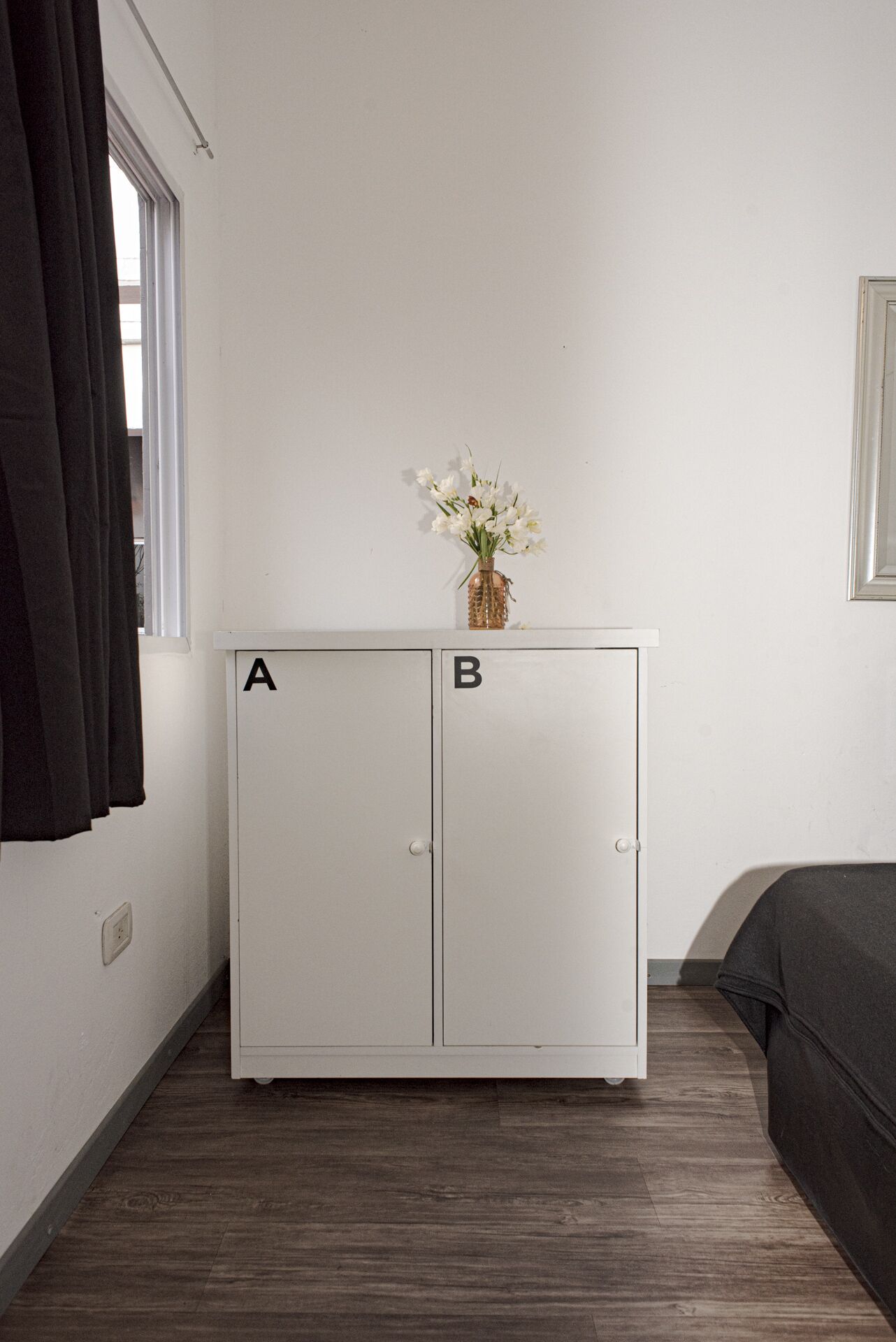 A small room with a white wardrobe labeled A and B, a window with curtains, a vase with flowers, and a bed with a dark cover.