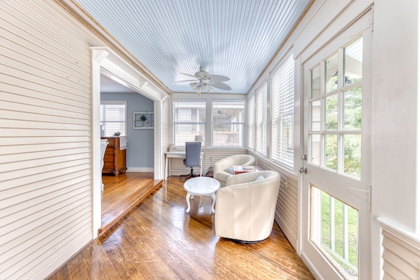 A sunroom with a ceiling fan, large windows, a wooden floor, and a seating area with a table and chairs.