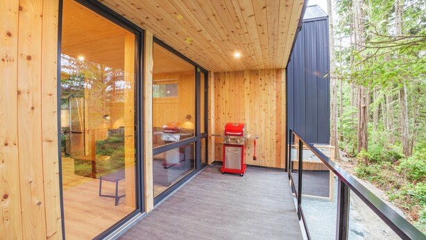 En moderne træhytte med store glasdøre, en terrasse og en rød grill.