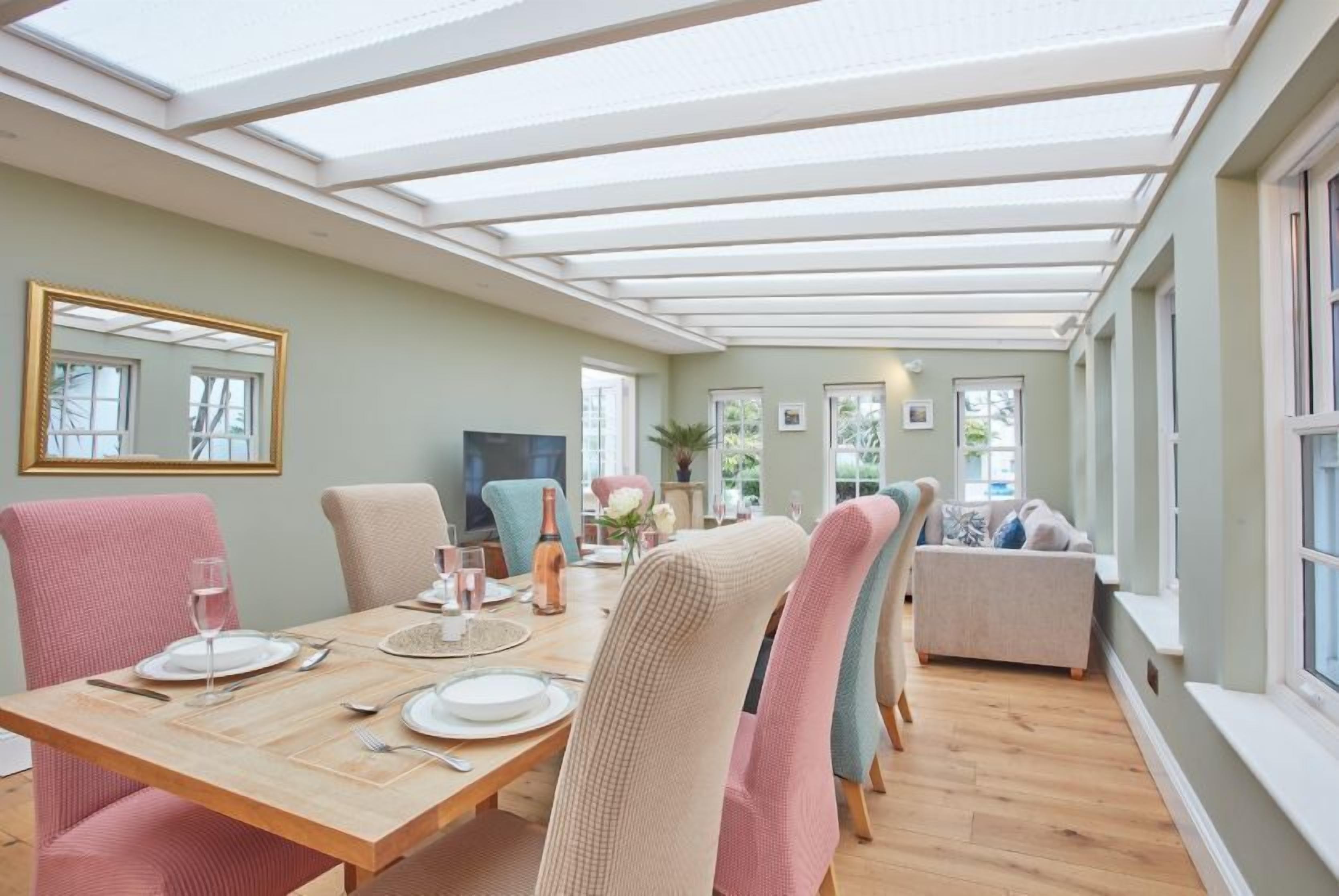 A dining area with a wooden table set for four, surrounded by chairs with a checkered pattern. A large mirror hangs on the wall, and there is a sofa in the background.