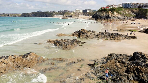 A coastal beach with rocky areas, a sandy shore, and buildings in the background.