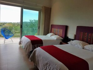 Two beds with red and white bedspreads, a balcony with a chair, and a sliding glass door offering a view of greenery and the sky.
